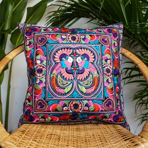 May include: A colorful embroidered pillow with a floral and bird design. The pillow is black with pink, blue, green, and yellow embroidery. The pillow is sitting on a wicker chair.