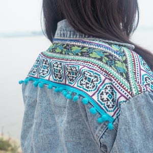 Peut inclure: Veste en jean avec un empiècement brodé à motifs géométriques en turquoise, blanc et violet. La veste est ornée de pompons turquoise le long des épaules et des manches. L'arrière-plan est une vue floue de l'eau et du ciel.