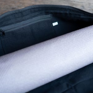 May include: A black canvas yoga bag with a zippered pocket on the inside. The bag is open and a rolled-up yoga mat is inside.