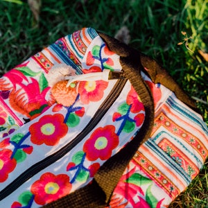 Puede incluir: Una bolsa de tela blanca con flores y rayas bordadas de colores. La bolsa tiene una cremallera y una correa negra.