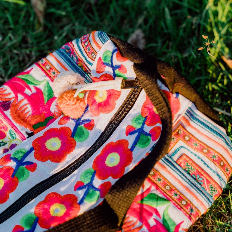 Puede incluir: Una bolsa de tela blanca con flores y rayas bordadas de colores. La bolsa tiene una cremallera y una correa negra.
