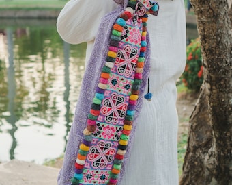 May include: A purple yoga mat bag with a colorful embroidered design and pom-poms. The bag is being carried by a person wearing a white dress.