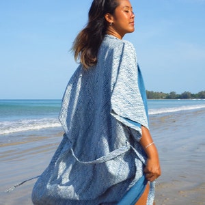 May include: A woman wearing a blue and white patterned kimono-style robe on a beach. The robe is tied at the waist and the fabric is flowing in the wind.