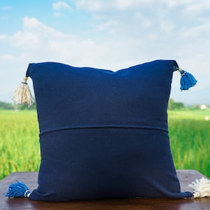 May include: A dark blue throw pillow with white and blue tassels. The pillow is sitting on a wooden surface with a green field in the background.