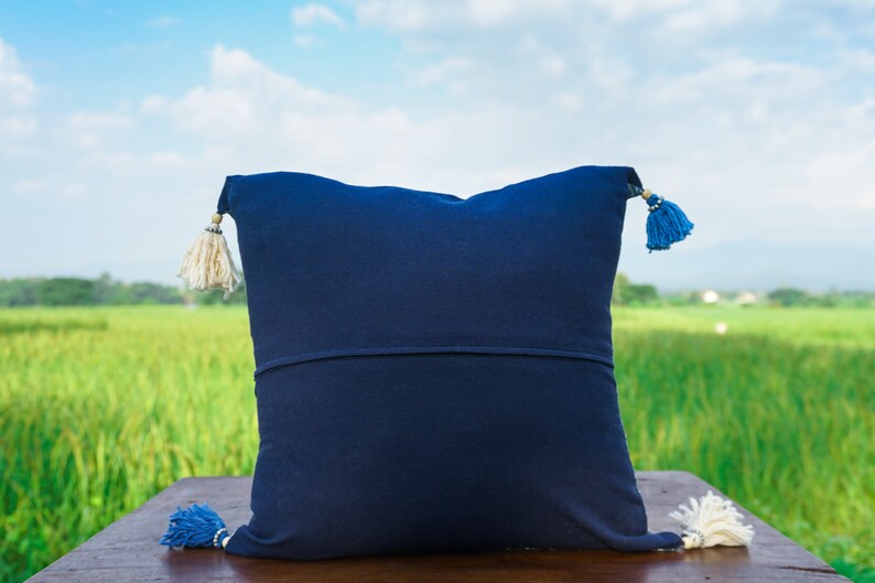 May include: A dark blue throw pillow with white and blue tassels. The pillow is sitting on a wooden surface with a green field in the background.