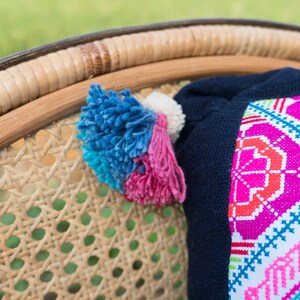 May include: A close-up of a woven wicker chair with a colorful embroidered pillow. The pillow has a navy blue backing and a white front with pink, red, blue, and green geometric patterns. The chair has a natural brown color.