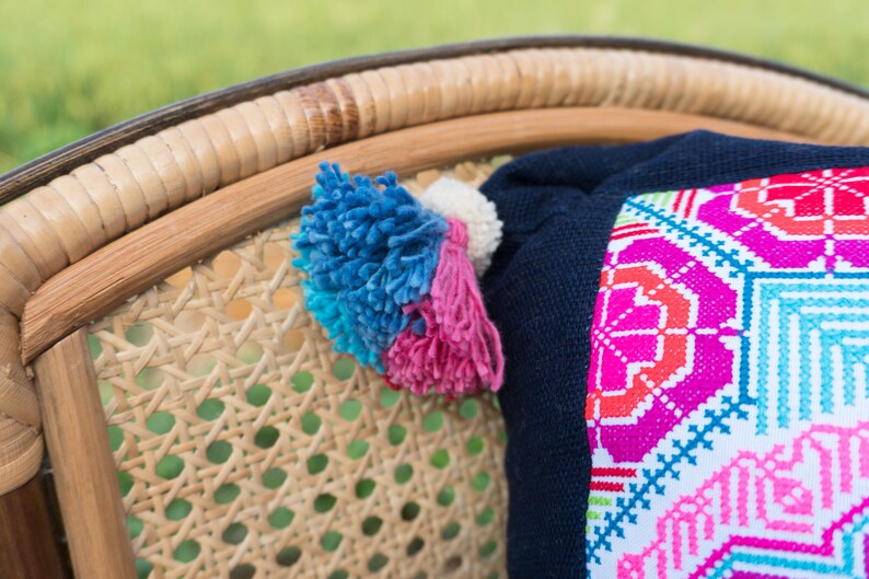 May include: A close-up of a woven wicker chair with a colorful embroidered pillow. The pillow has a navy blue backing and a white front with pink, red, blue, and green geometric patterns. The chair has a natural brown color.
