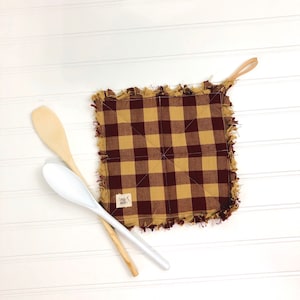 May include: A brown and tan checkered pot holder with a frayed edge and a small brown tag that says "Love, Made." The pot holder has a loop for hanging.