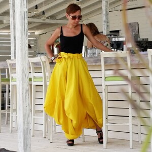 May include: A woman wearing a black tank top and a bright yellow skirt with a high waist and a large bow. She is wearing black sandals. The background includes white chairs and a bar area.