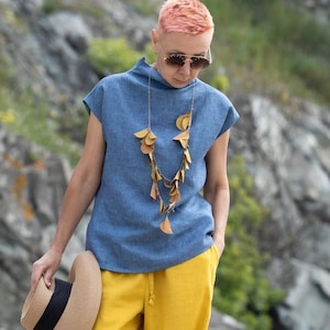 A woman with short hair walks along the beach. She carries a straw hat in her hand. She is dressed in a beautiful linen blouse with short sleeves and wide trousers. Behind her are rocks.