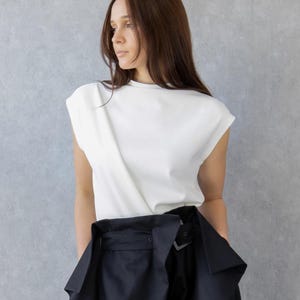 May include: A woman wearing a white sleeveless top and black high-waisted pants. The pants have a unique design with large pockets and a belt. She is also wearing black boots. The background is a plain gray wall.