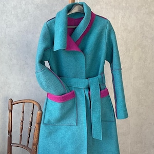 May include: A teal coat with magenta trim and a matching belt hangs on a wooden coat rack. The coat features a shawl collar, pockets, and a long, knee-length design. A wooden chair sits to the left of the coat.