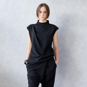 May include: A woman wearing a black outfit. The ensemble includes a sleeveless top with a high neckline and draped pants. She is also wearing black boots and a black bracelet. The outfit has a modern, minimalist style.