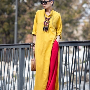May include: A woman wearing a mustard yellow tunic dress with a high side slit, paired with bright red pants. She is accessorized with a statement necklace, sunglasses, and a woven handbag. The outfit is a bold mix of colors and textures.