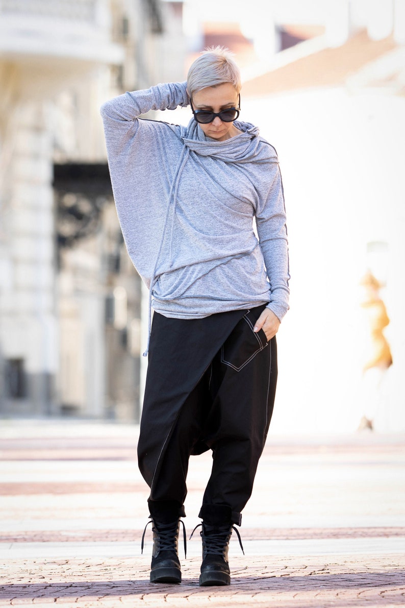 May include: A woman wearing a gray hooded sweater with a cowl neck and black pants with a unique design. She is wearing black boots and sunglasses.