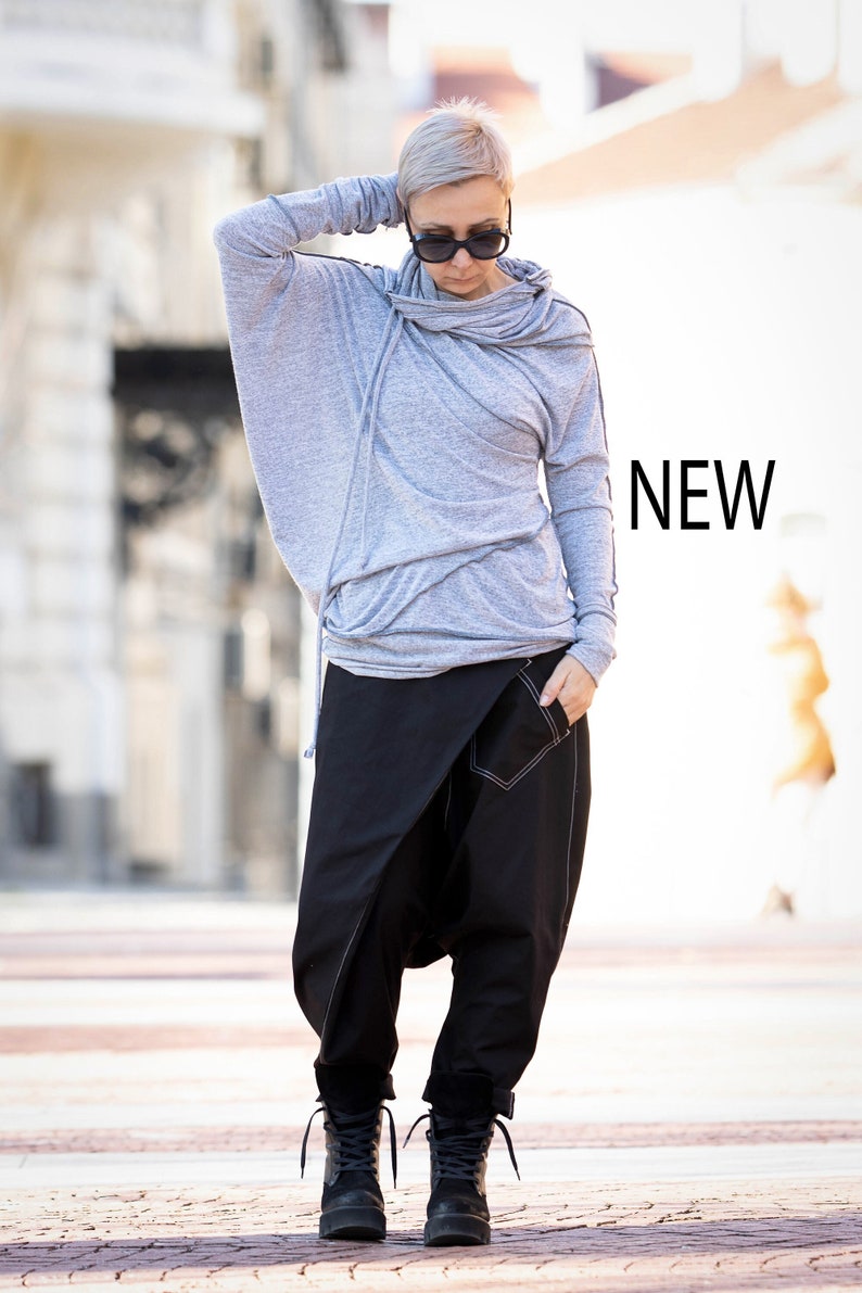 May include: A gray hooded sweatshirt with a draped neckline and long sleeves. The model is wearing black pants with a unique design and black boots. The word "NEW" is visible in the image.
