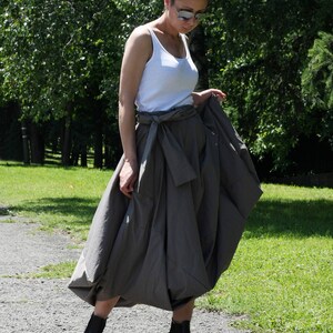 May include: A woman wearing a white tank top, a gray skirt with a tie waist, and black wedge sandals with white soles. The woman is wearing sunglasses and standing on a paved path in a park.