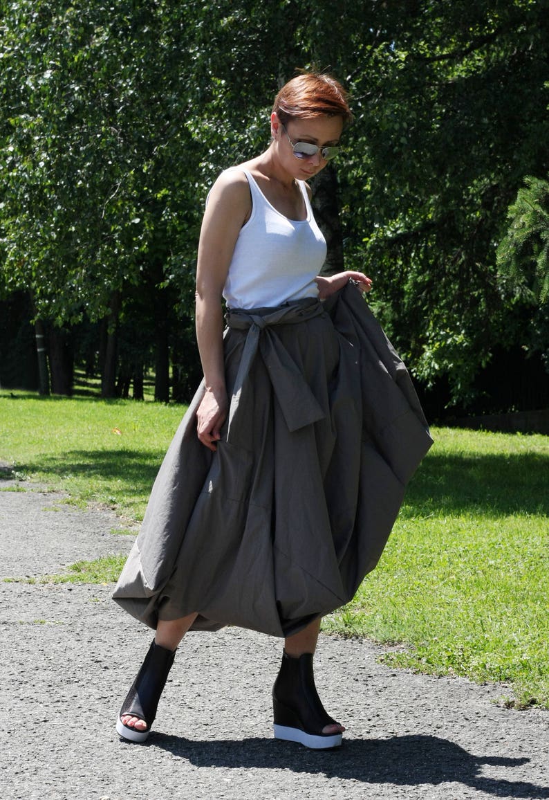 May include: A woman wearing a white tank top, a gray skirt with a tie waist, and black wedge sandals with white soles. The woman is wearing sunglasses and standing on a paved path in a park.