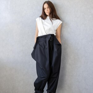 May include: A woman wearing a white sleeveless top and black high-waisted pants with a unique wrap design. The pants have a wide leg and are paired with black platform boots. The background is a plain gray wall.