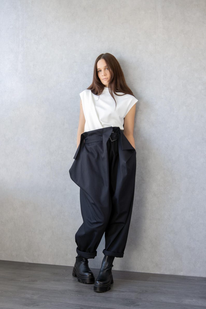 May include: A woman wearing a white sleeveless top and black high-waisted pants with a unique wrap design. The pants have a wide leg and are paired with black platform boots. The background is a plain gray wall.