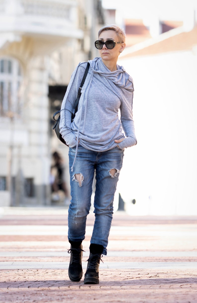 May include: A woman wearing a gray hooded sweatshirt, ripped blue jeans, and black ankle boots. She is walking on a brick pathway.