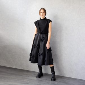 May include: A black outfit consisting of a sleeveless top and a midi skirt. The skirt features multiple bow accents. The model is wearing black boots and socks. The background is a light gray wall and a gray floor.