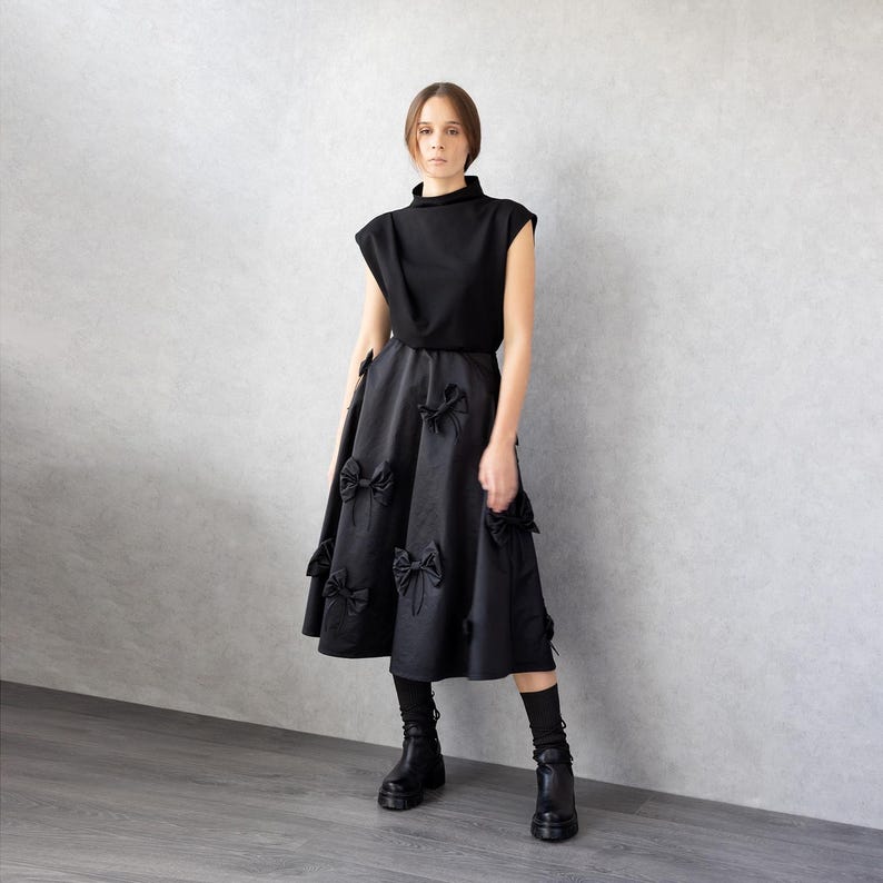 May include: A black outfit consisting of a sleeveless top and a midi skirt. The skirt features multiple bow accents. The model is wearing black boots and socks. The background is a light gray wall and a gray floor.