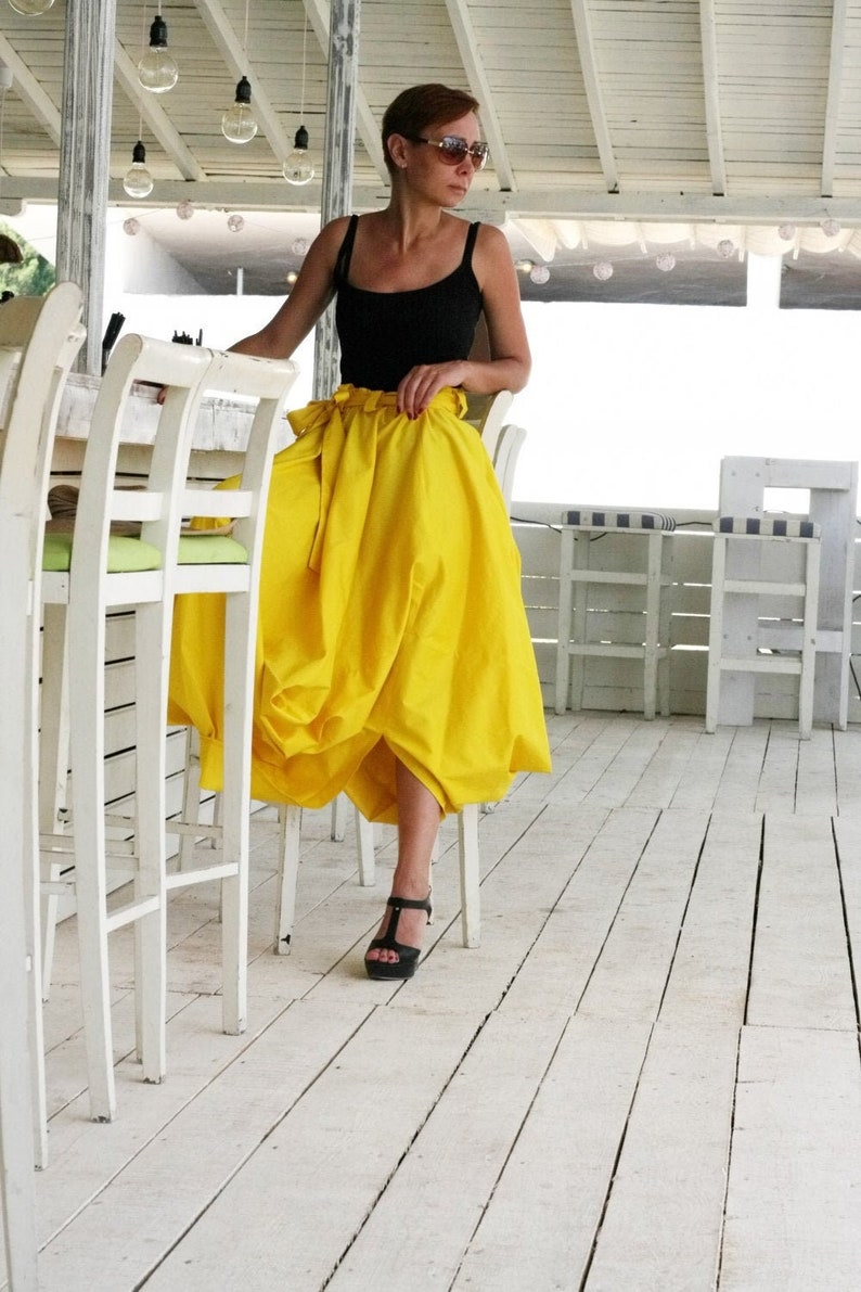 May include: A woman wearing a black tank top and a bright yellow skirt. The skirt has a unique, gathered design. She is wearing black sandals. The setting appears to be a white-painted outdoor patio with hanging lights.