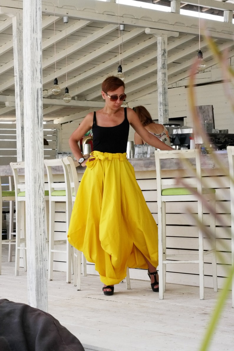 May include: A woman wearing a black tank top and a bright yellow skirt with a high waist and a large bow. She is wearing black sandals. The background includes white chairs and a bar area.