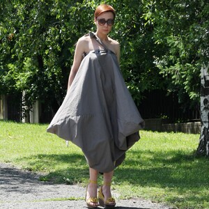 May include: A woman wearing a gray, asymmetrical dress with a unique, voluminous silhouette. The dress has a single shoulder strap and a flowing, draped design. She is wearing yellow platform sandals with floral accents and sunglasses.