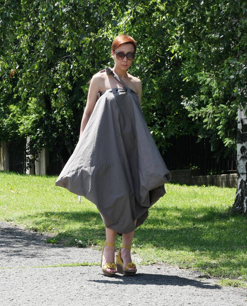 May include: A woman wearing a gray, asymmetrical dress with a unique, voluminous silhouette. The dress has a single shoulder strap and a flowing, draped design. She is wearing yellow platform sandals with floral accents and sunglasses.