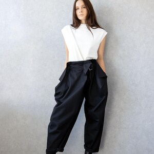 May include: A woman wearing a white sleeveless top and black high-waisted pants. The pants have a unique design with a belt and large pockets. She is also wearing black boots with a chunky sole.