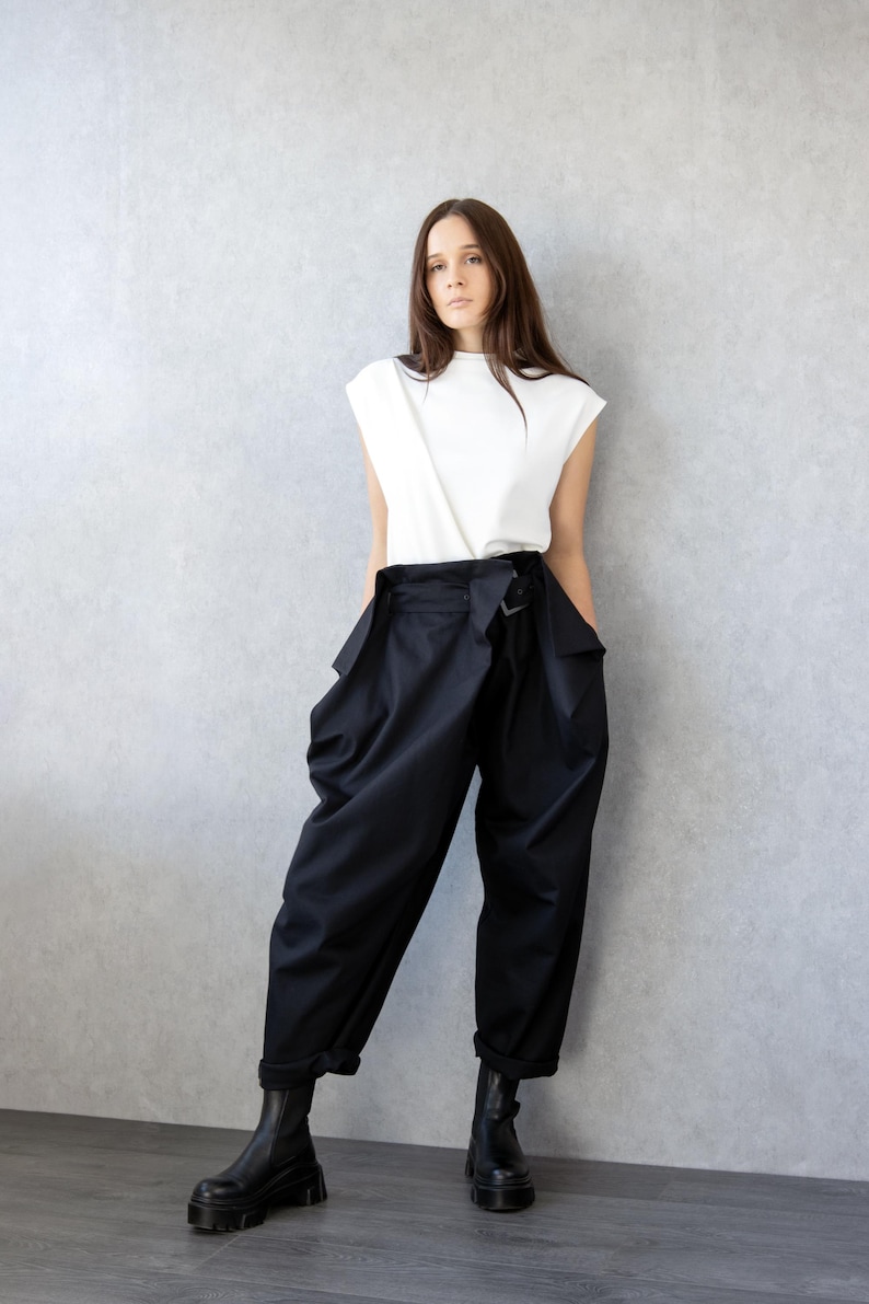 May include: A woman wearing a white sleeveless top and black high-waisted pants. The pants have a unique design with a belt and large pockets. She is also wearing black boots with a chunky sole.