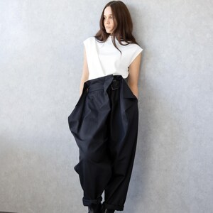 May include: A woman's outfit featuring a white sleeveless top and high-waisted, wide-leg black pants with a belt. The pants have a unique, voluminous design. Black ankle boots complete the look. The outfit is modern and stylish.