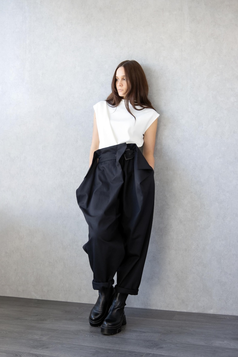 May include: A woman's outfit featuring a white sleeveless top and high-waisted, wide-leg black pants with a belt. The pants have a unique, voluminous design. Black ankle boots complete the look. The outfit is modern and stylish.