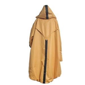 Op de afbeelding: Een mosterdgele cape met capuchon en een opvallende zwarte verticale streep die over het midden van de rug en de capuchon loopt. Het kledingstuk heeft een wijd, los silhouet met een V-vormig rugpand en lange mouwen, gepresenteerd tegen een witte achtergrond.