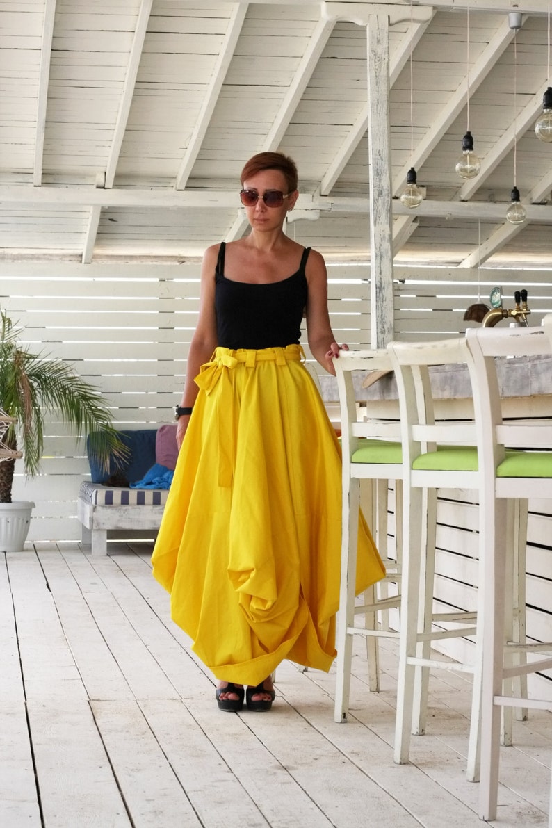 May include: A woman wearing a black tank top and a bright yellow skirt with a matching belt. The skirt has a unique, draped design. She is wearing sunglasses and black sandals. The background includes a white wooden structure and bar stools.