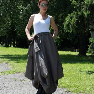 May include: A woman wearing a white tank top, a gray skirt with a wrap-around tie, and black sandals with white soles. The outfit is worn outdoors on a sunny day, with a background of green trees and a paved path.