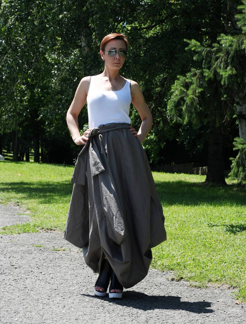 May include: A woman wearing a white tank top, a gray skirt with a wrap-around tie, and black sandals with white soles. The outfit is worn outdoors on a sunny day, with a background of green trees and a paved path.