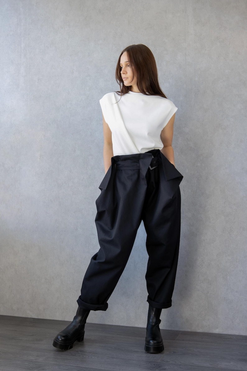 May include: A woman wearing a white sleeveless top and black high-waisted pants. The pants have a unique design with large pockets and a belt. She is also wearing black boots. The background is a plain gray wall.