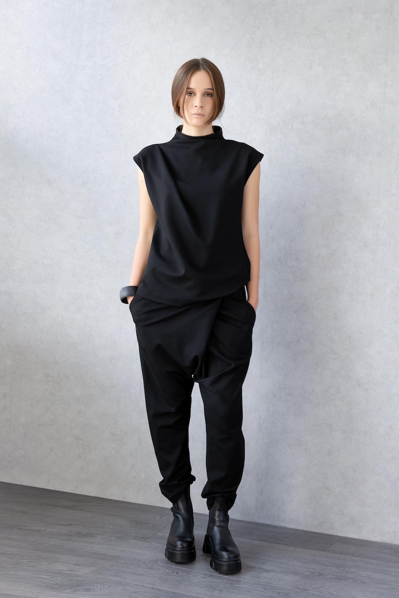 May include: A black ensemble featuring a mock neck top and drop-crotch pants. The top has short sleeves, and the pants have a unique draped design. The outfit is paired with black boots and a black bracelet. The clothing is made of a smooth fabric.