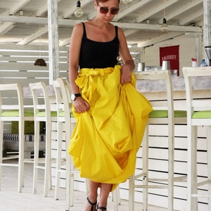 May include: A woman wearing a black tank top and a bright yellow skirt. She is wearing black sandals and sunglasses. The background includes a white bar with chairs and hanging light bulbs.