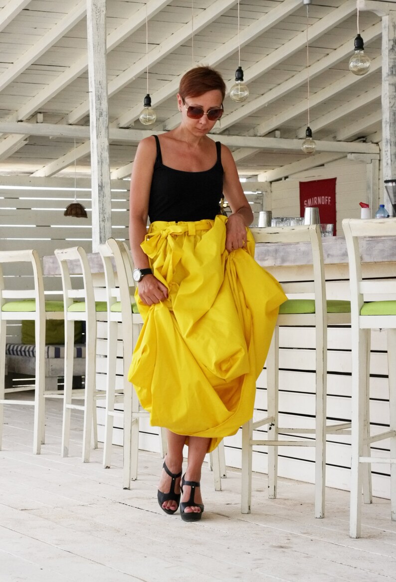 May include: A woman wearing a black tank top and a bright yellow skirt. She is wearing black sandals and sunglasses. The background includes a white bar with chairs and hanging light bulbs.
