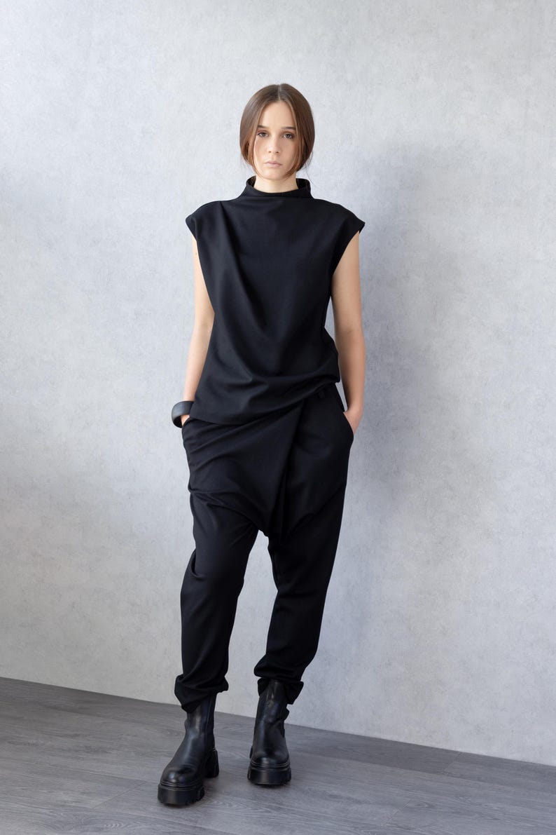 May include: A woman wearing a black outfit. The ensemble includes a sleeveless top with a high neckline and draped pants. She is also wearing black boots and a black bracelet. The outfit has a modern, minimalist style.