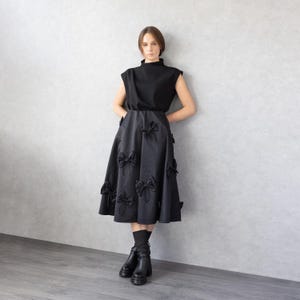 May include: A black sleeveless top and a black midi skirt adorned with numerous bow details. The outfit is paired with black boots. The model is standing against a gray wall.