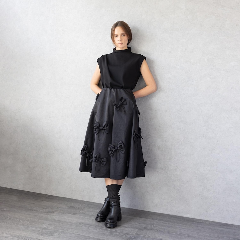 May include: A black sleeveless top and a black midi skirt adorned with numerous bow details. The outfit is paired with black boots. The model is standing against a gray wall.