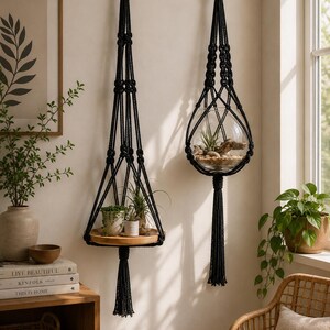 Peut inclure: Deux suspensions murales en macramé noir avec anneaux et pompons en bois. L'une supporte une étagère en bois avec de petites plantes en pot, l'autre un terrarium en verre avec des plantes et des pierres.
