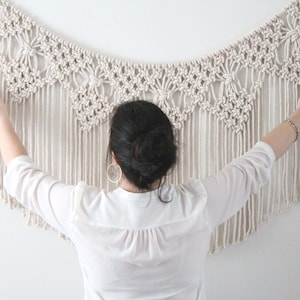 May include: A cream-coloured macrame wall hanging with detailed knotting and fringe. The textile is displayed against a white wall. The macrame has a scalloped design along the top edge.