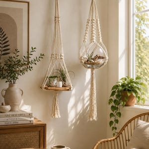 May include: Two cream-coloured macrame plant hangers, one with a wooden shelf holding small potted plants, the other a glass terrarium. The room has a neutral colour palette and natural light.