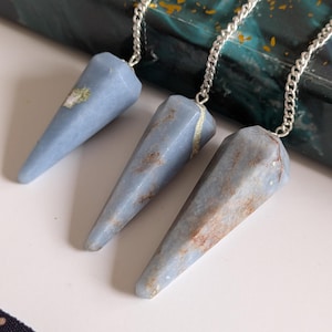 May include: Three blue crystal pendulums with silver chains. Each pendulum has a faceted, conical shape. The crystals have a pale blue hue with natural variations of colour, including brown and white markings. The chains are made of small, linked metal pieces.